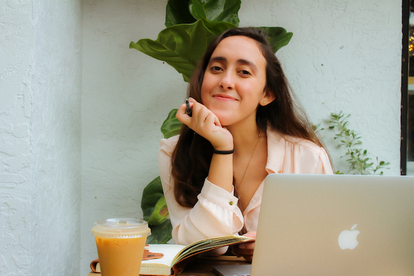 a girl with her chin in her hand, sitting in front of a computer, notebook, and coffee.