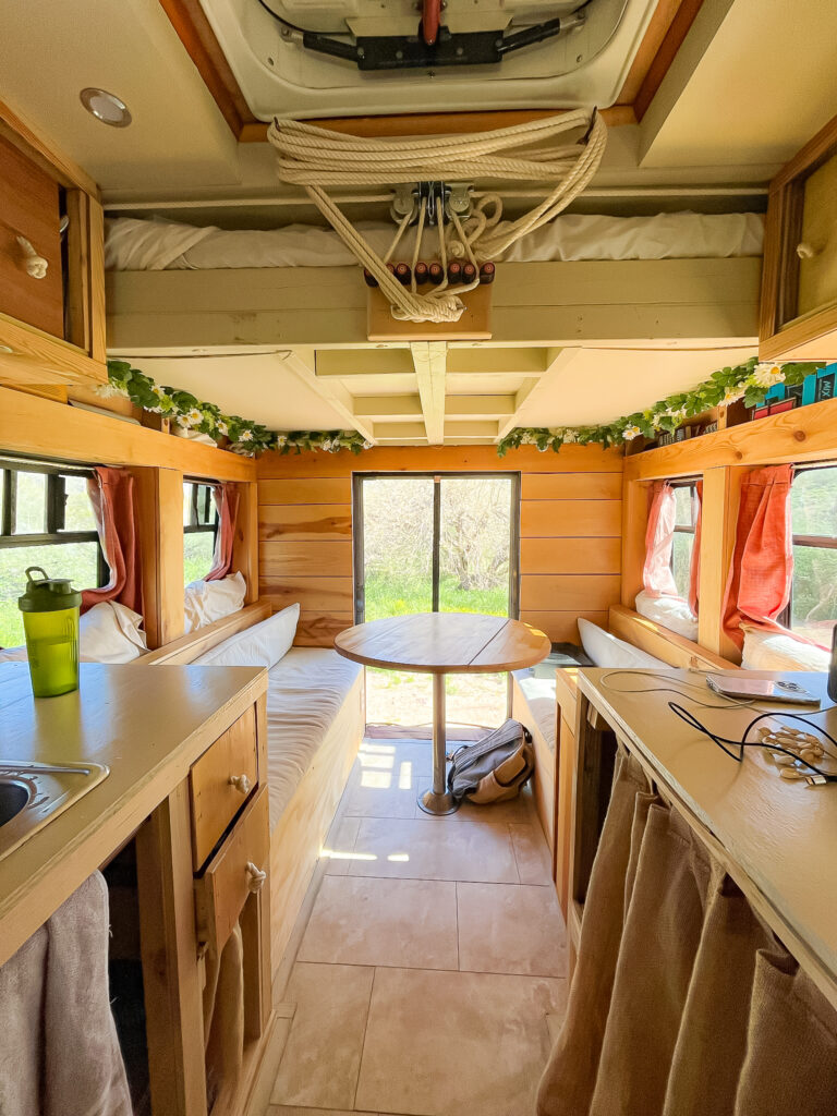 the inside of a converted shuttle bus. The back door of the bus is open, it is behind a round wooden table and two benches on each side of the table. A bed is on pulleys against the ceiling.